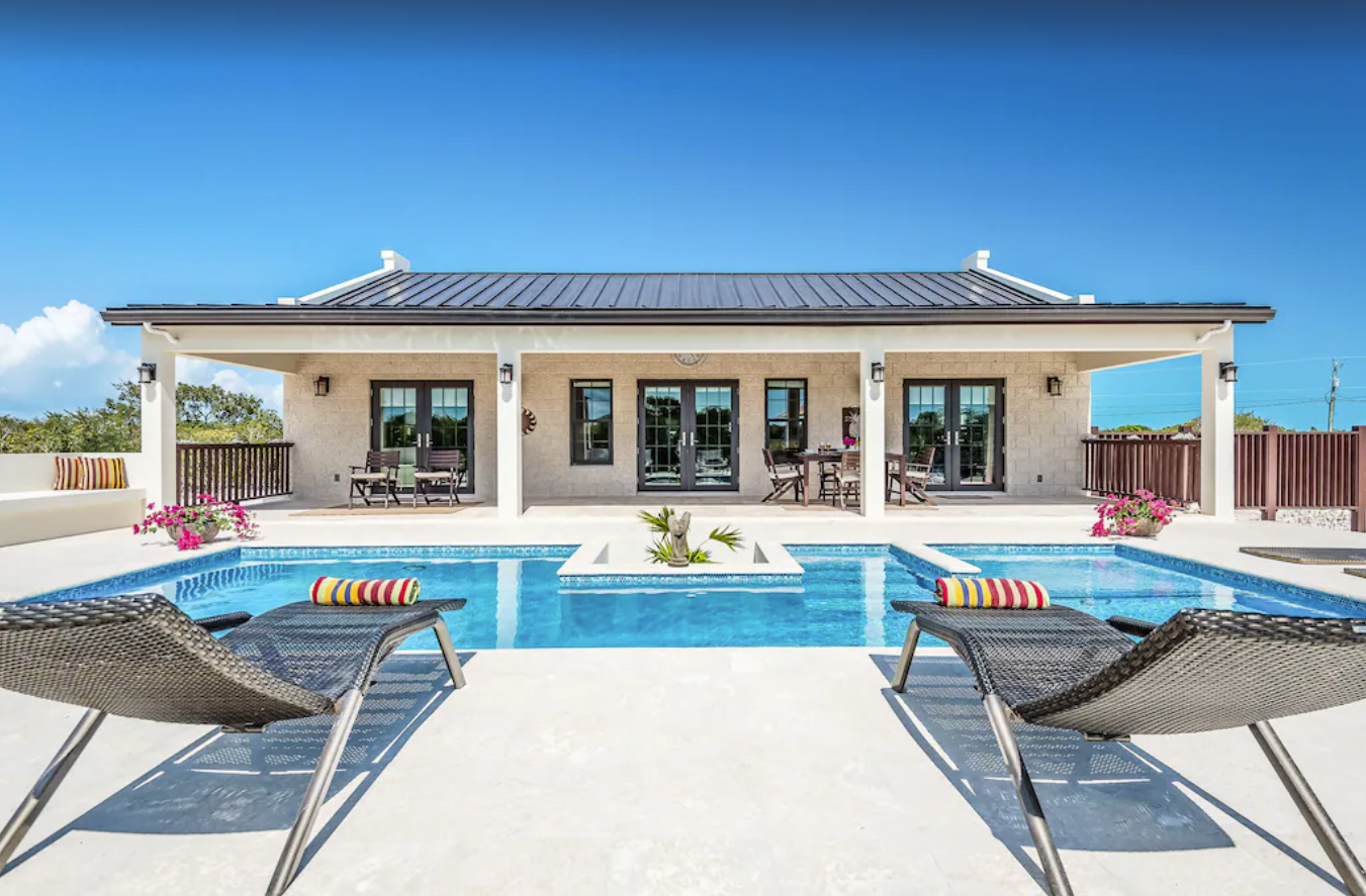 An exterior image of a small two-bedroom villa in Turks and Caicos. The private backyard pool area has lounge chairs and is surrounded by a privacy fence.