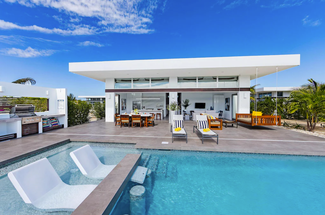 The private backyard pool area of a modern Turks and Caicos rental villa features a large pool, lounge chairs, an outdoor dining area and grilling area, and privacy fencing.