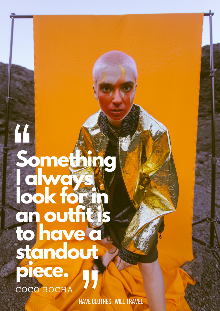 A model stands in front of a bright orange background. They're wearing a gold foil jacket and has orange and yellow flames painted on their face. White text over the image says, "Something I always look for in an outfit is to have a standout piece. - Coco Rocha"