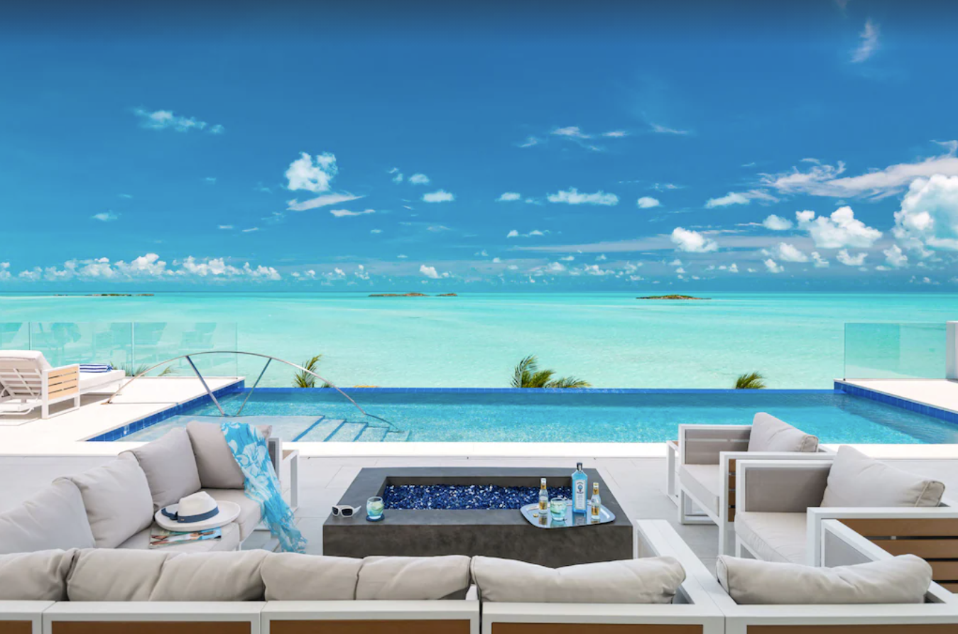 The view of the crystal blue ocean and clear skies from the private backyard pool and patio of a Turks and Caicos villa. The patio includes comfortable couches and chairs around a firepit, tanning chairs, and a large infinity pool.