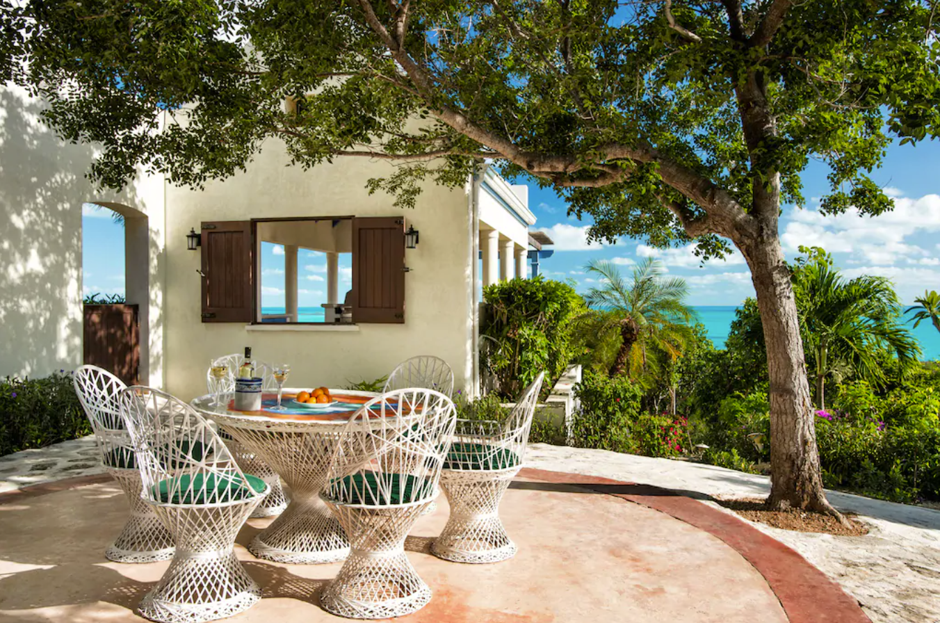 This Turks and Caicos villa features an outdoor eating area, furnished with chairs and a table shaded by a tropical tree, overlooking the crystal blue ocean.