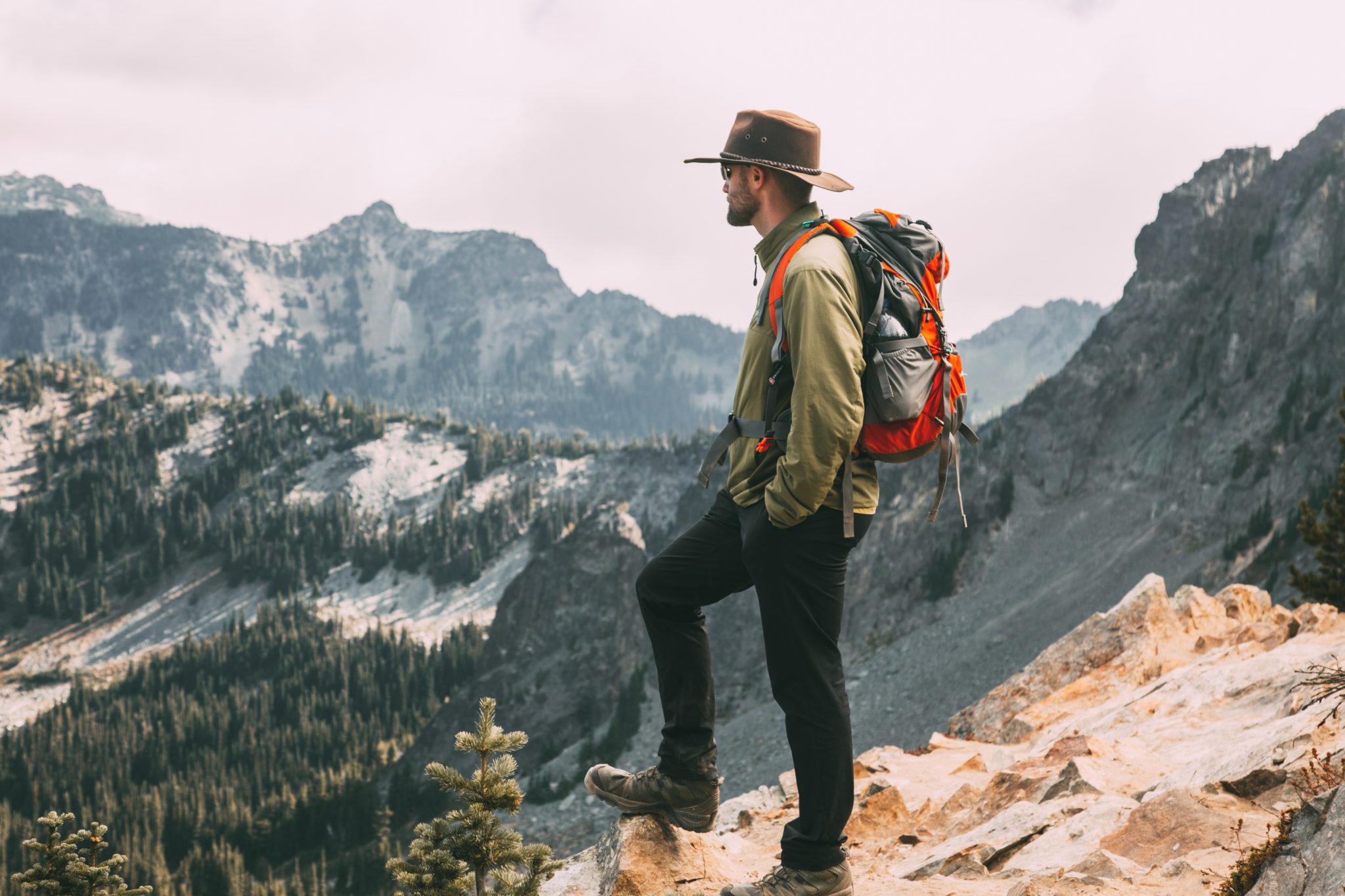 A man stands on a mountain range, overlooking the scenery. He's wearing a hiking jacket and pants, hat, and hiking pack on his pack while he looks out over the valleys of trees and mountains.