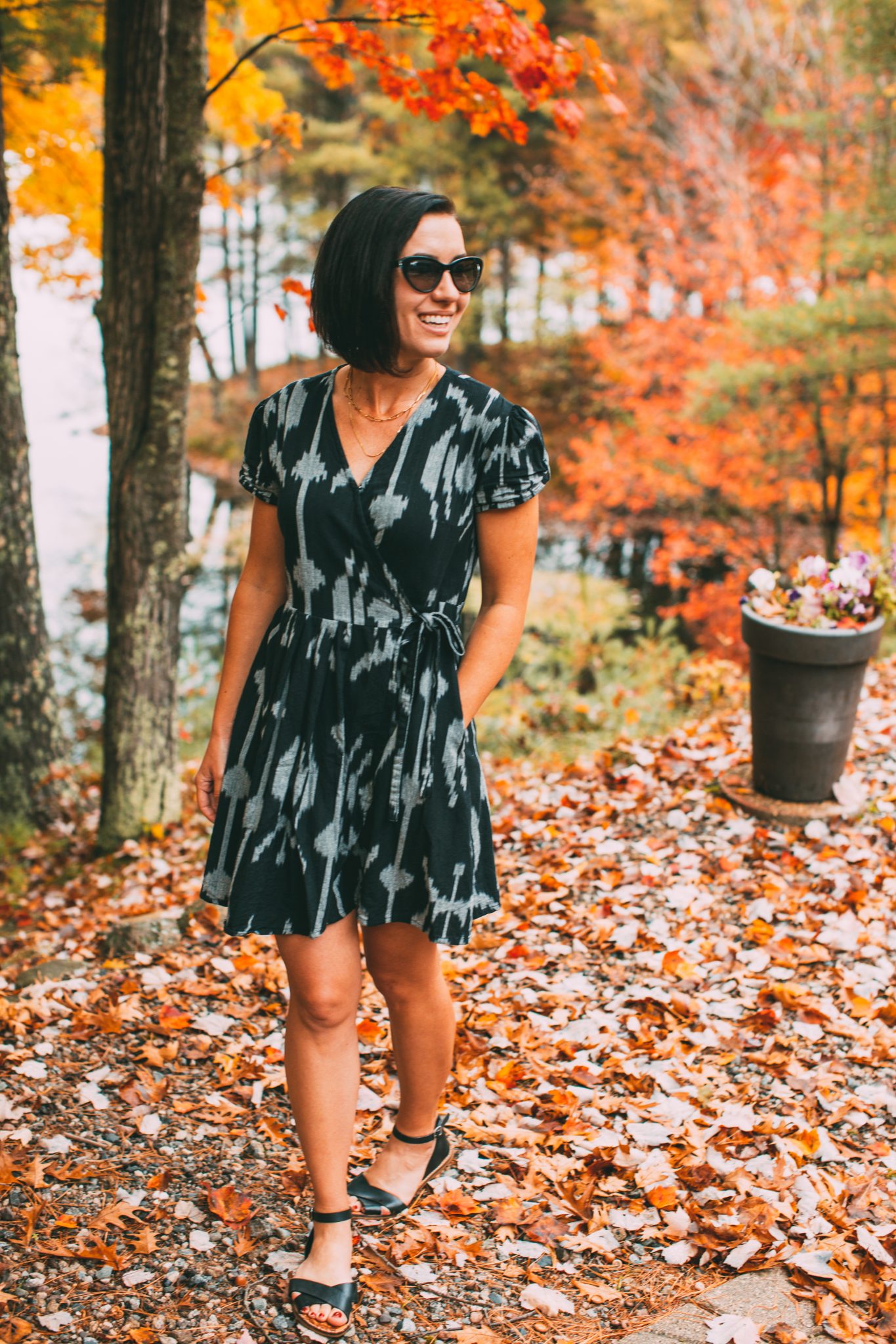 A woman poses in a picturesque fall landscape with colorful trees and fallen leaves behind her. She models a black and gray patterned Passion Lillie dress.