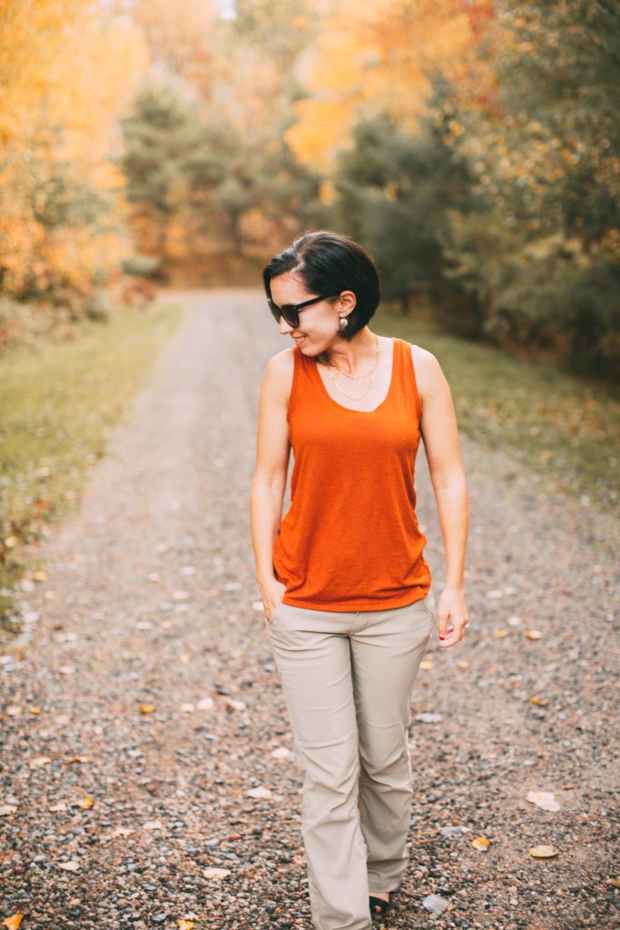 A woman walks along a dirt road wearing an orange tank top and khaki pants from the sustainable travel clothing brand prAna.