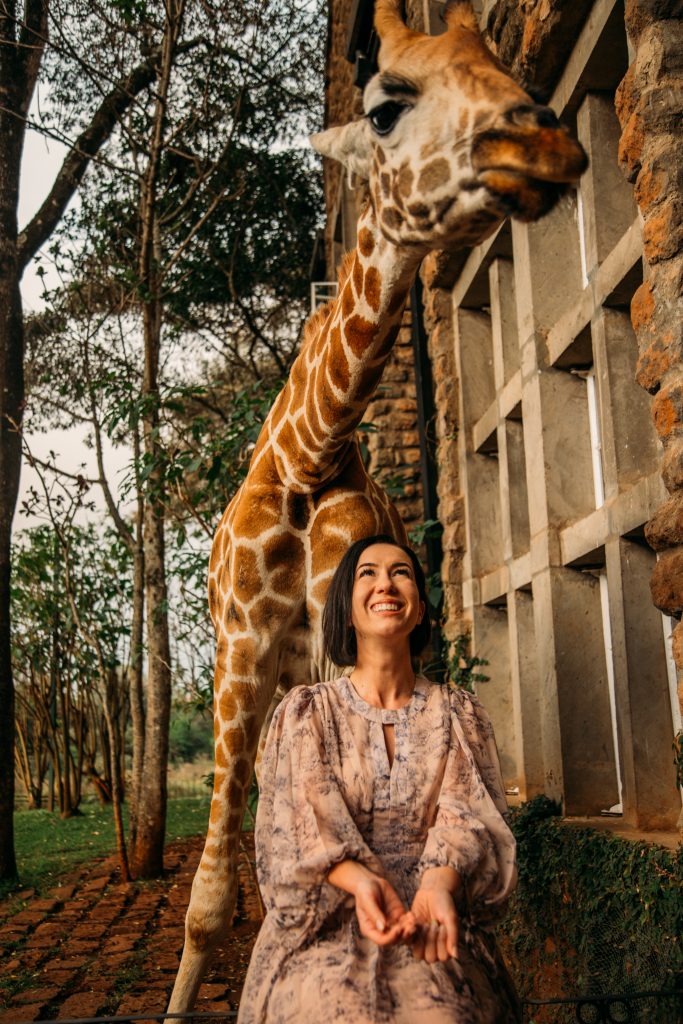 A woman poses outside, sitting in front of a giraffe that looms over her. The woman is wearing a light brown long sleeve dress with flowy sleeves and skirt, and she's smiling up at the giraffe.