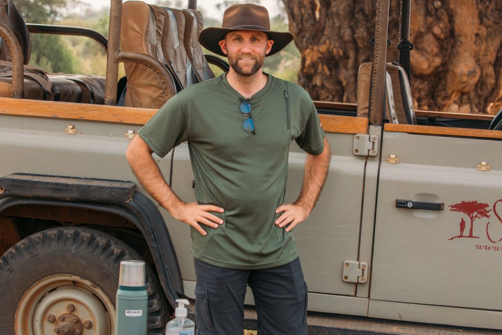 A man standing in front of a safari vehicle while wearing a SCOTTeVEST performance shirt in green and an oil skin hat.