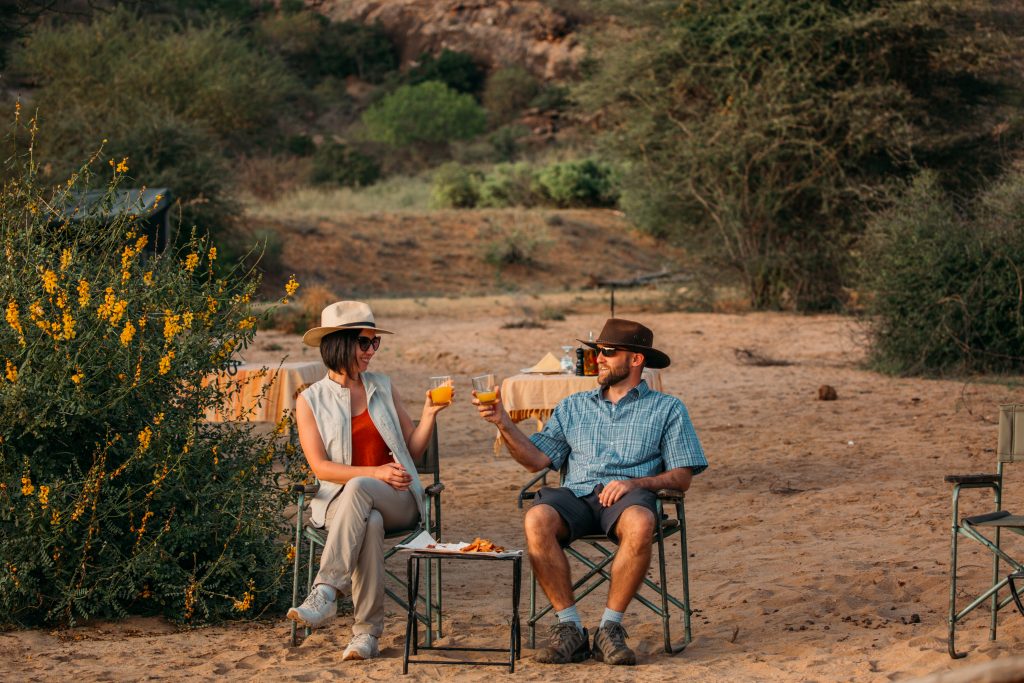 A man and woman sit on camping chairs at a camping site in the Kenyan desert. They're wearing outdoorsy clothing -- short-sleeve tops, hats, sunglasses, and light weight hiking pants, and cheersing their drinks.