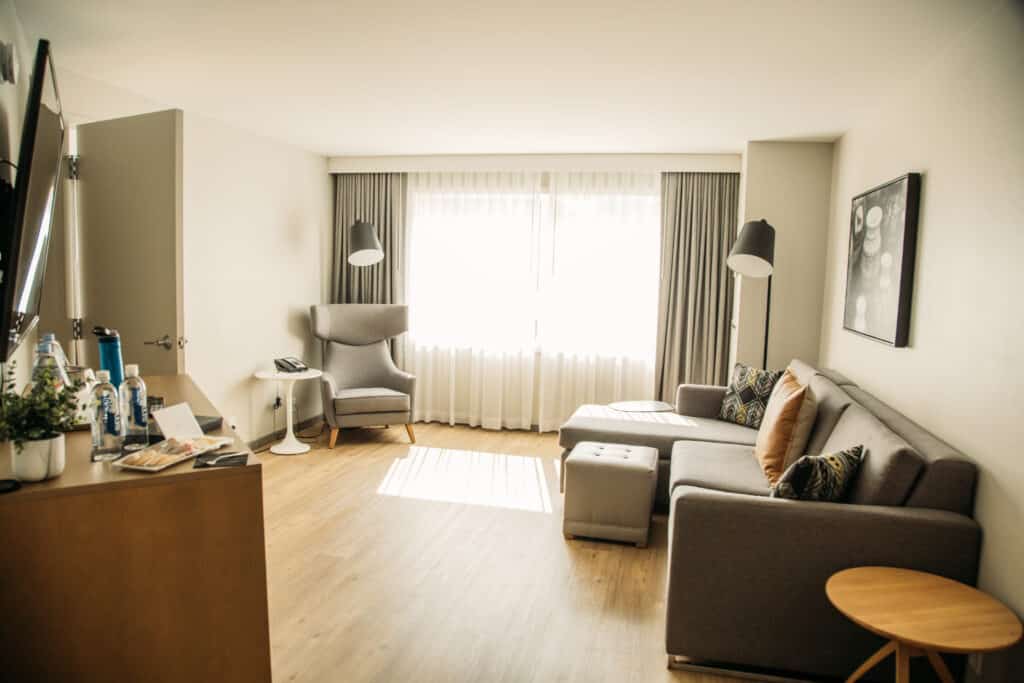 A living room features wooden floors and modern furnishings in shades of grey and beige. The room has off-white walls, black lamps, a white tulip-style telephone table next to a chair, and a couch with an ottoman. A desk in the foreground features two water bottles, snacks, a plant, and other amenities. 