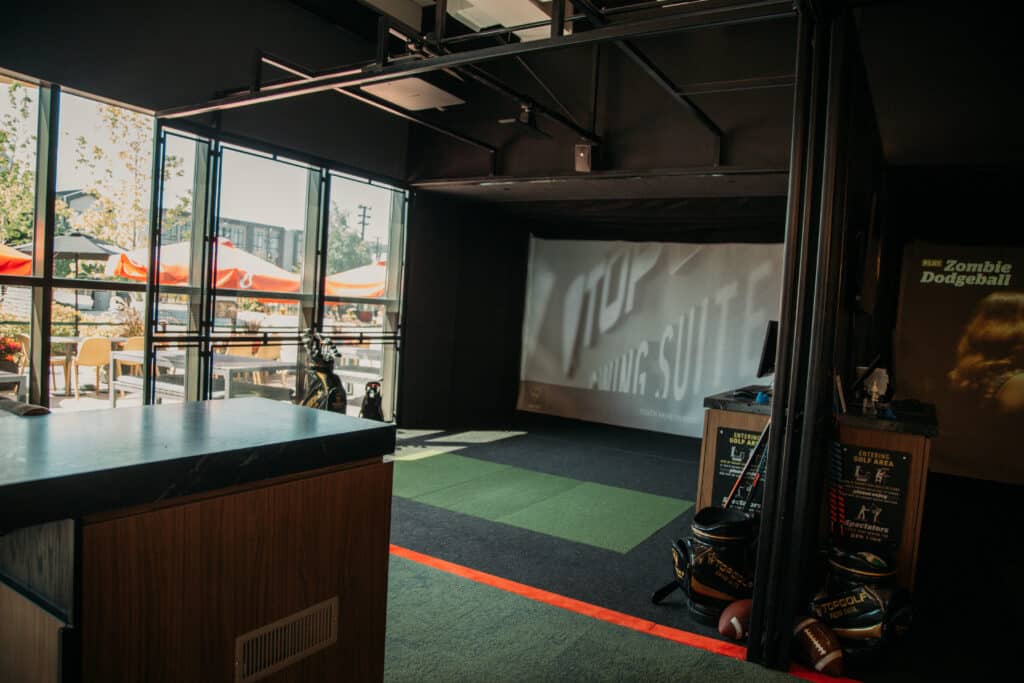 A virtual golf-swing suite is shown against a backdrop of windows. There is green AstroTurf on the floor and a screen against the wall. 