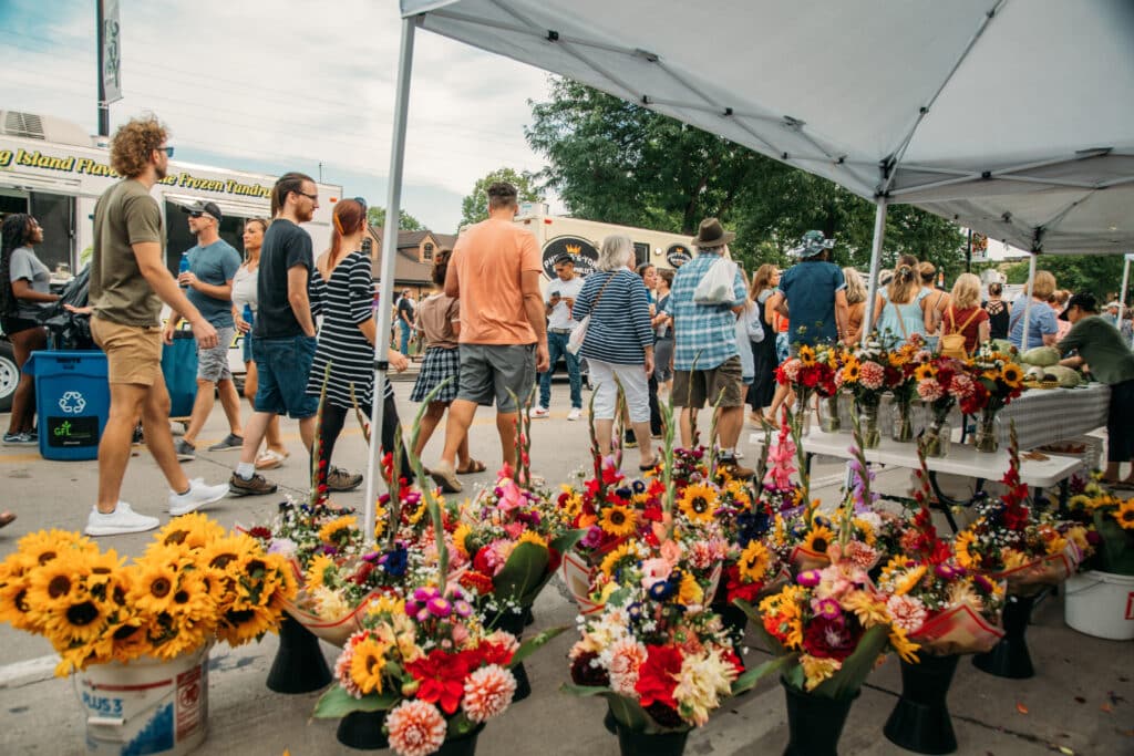 A crowd of people in summer attire filing past a flower booth at an outdoor farmers market. The booth features bouquets of sunflowers, dahlias, and lilies. Food trucks are set up in the background of the market. 