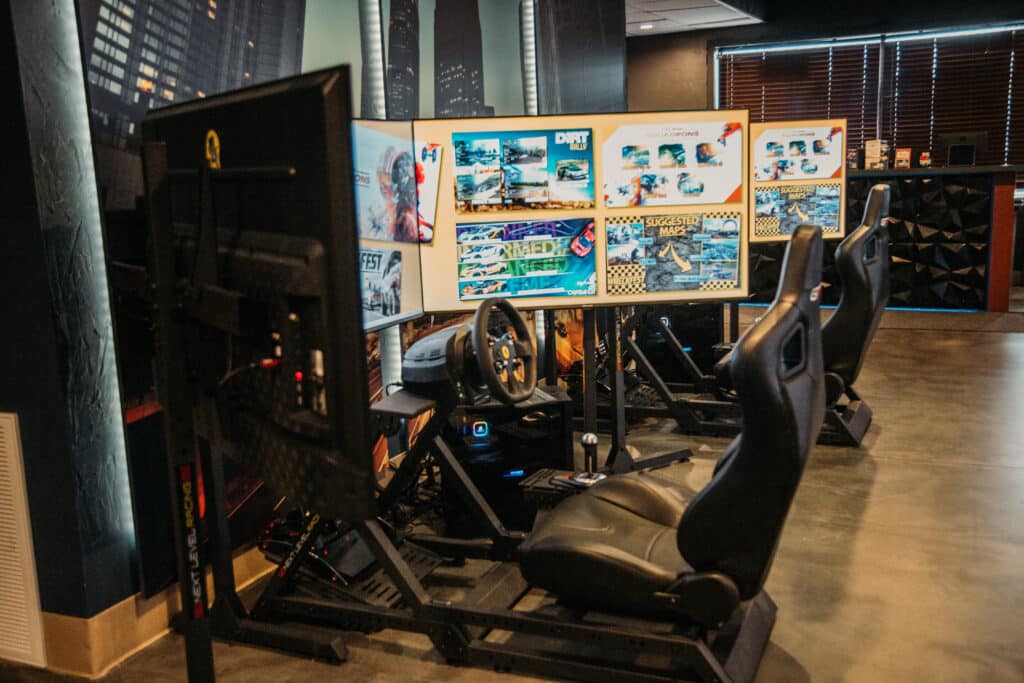 Two driving arcade rides are shown with black seats, steering wheels, and yellow digital screens. On the screens are many options and details for playing the driving video game. 