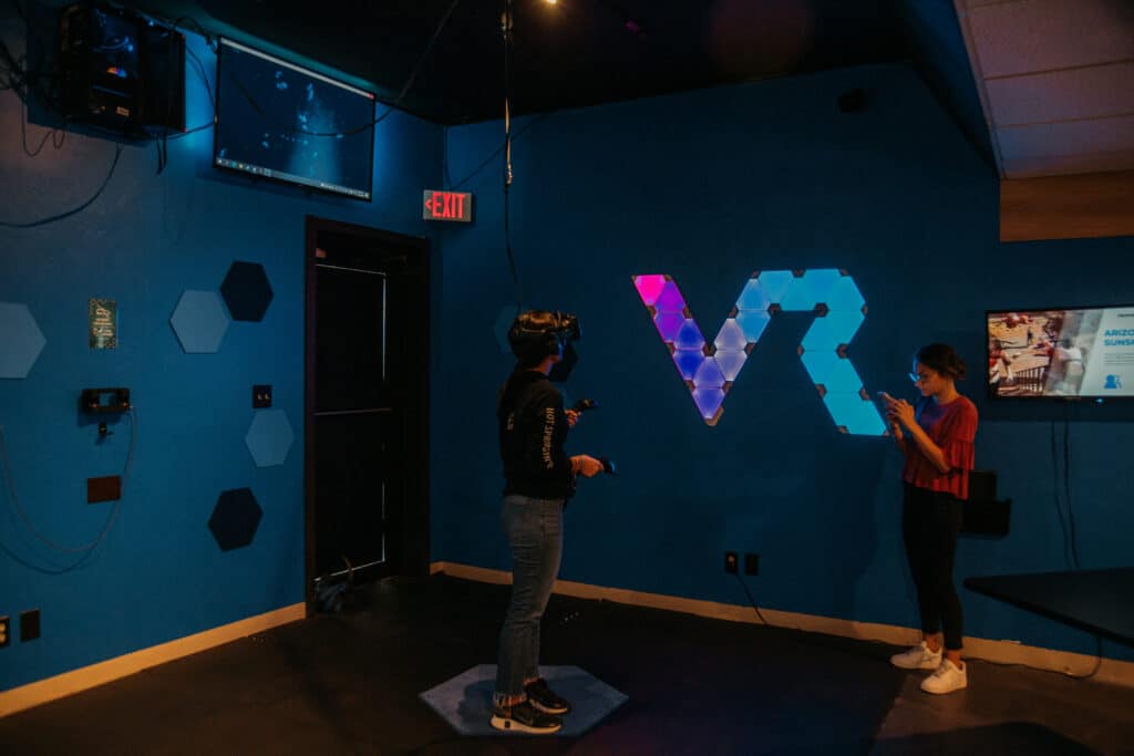 Two teens are in a dark room that says VR on the navy-blue wall. One is wearing a VR headset, while the other uses their phone to snap a photo. 