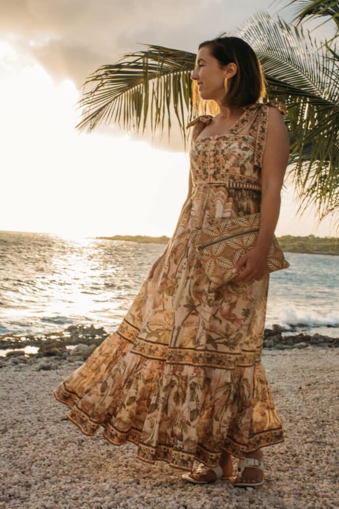 Lindsey of Have CLothes, Will Travel wearing a Zimmermann maxi dress, Carry Courage cork clutch, and Tory Burch sandals, standing on a rocky beach at sunset