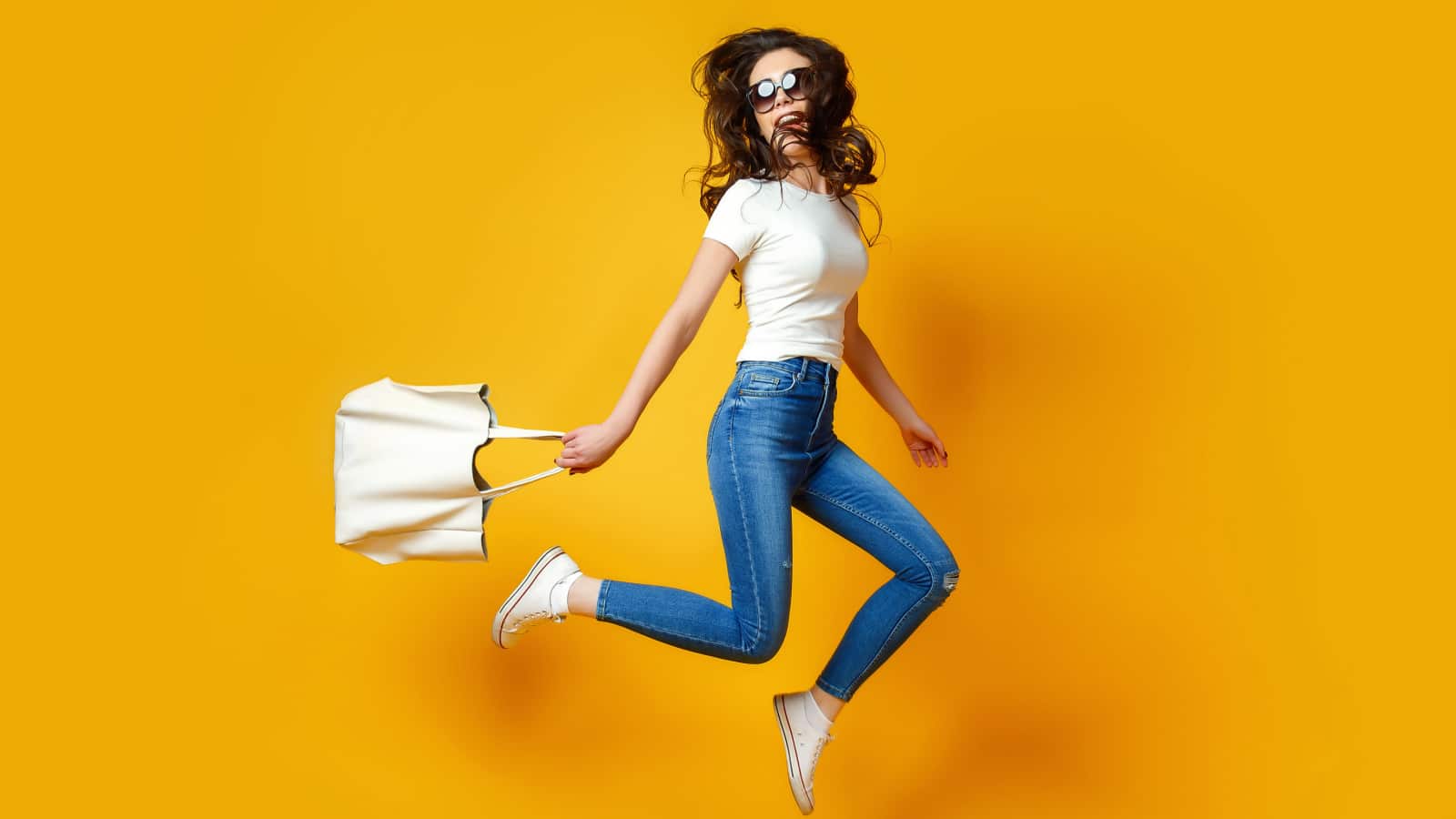 Beautiful young woman in sunglasses, white shirt, blue jeans posing, jumping with bag on the yellow background