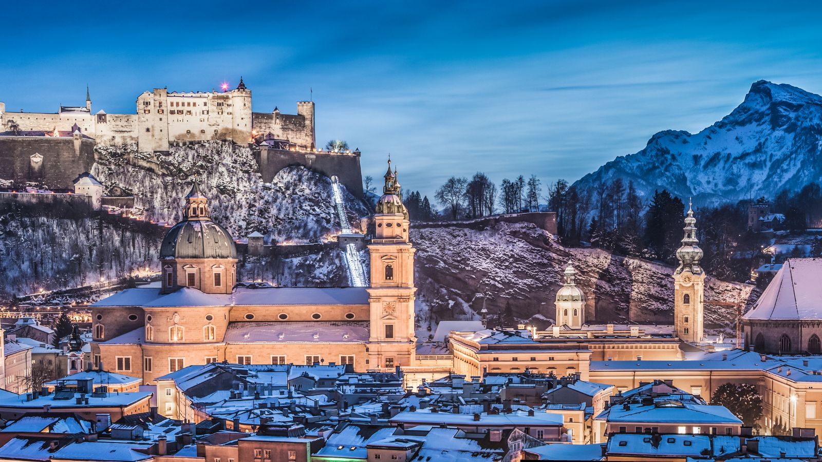 A snowy landscape image of Salzburg, Austria, showing a large castle at the top of a tall hill and the city buildings blow, covered in shadow and snow.