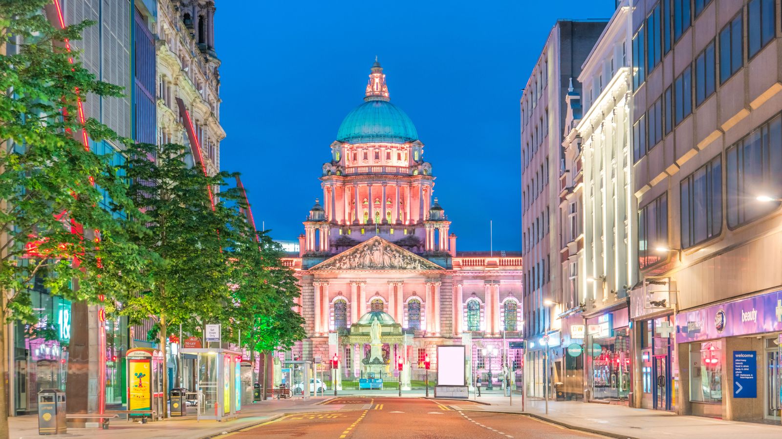 A view of Belfast City Hall at dusk in Belfast, Northern Ireland. The quiet street is lined with buildings leading towards the city center.