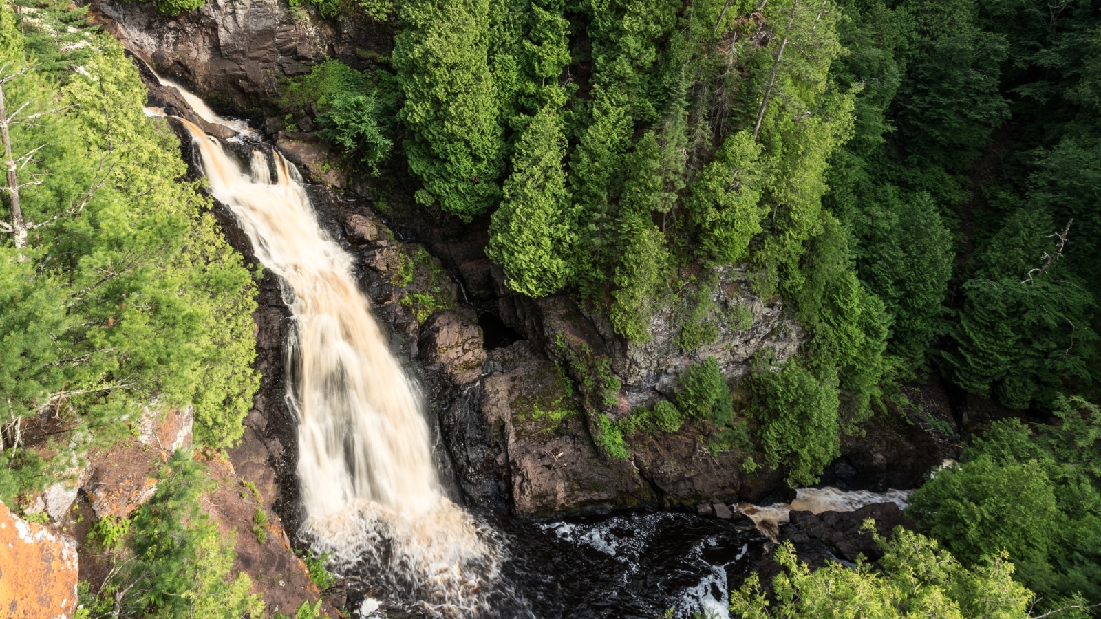 An aerial photo of Big Manitou Falls in Pattison State Park in Wisconsin. The giant waterfall flows down the mountainside, surrounded by lush green trees.