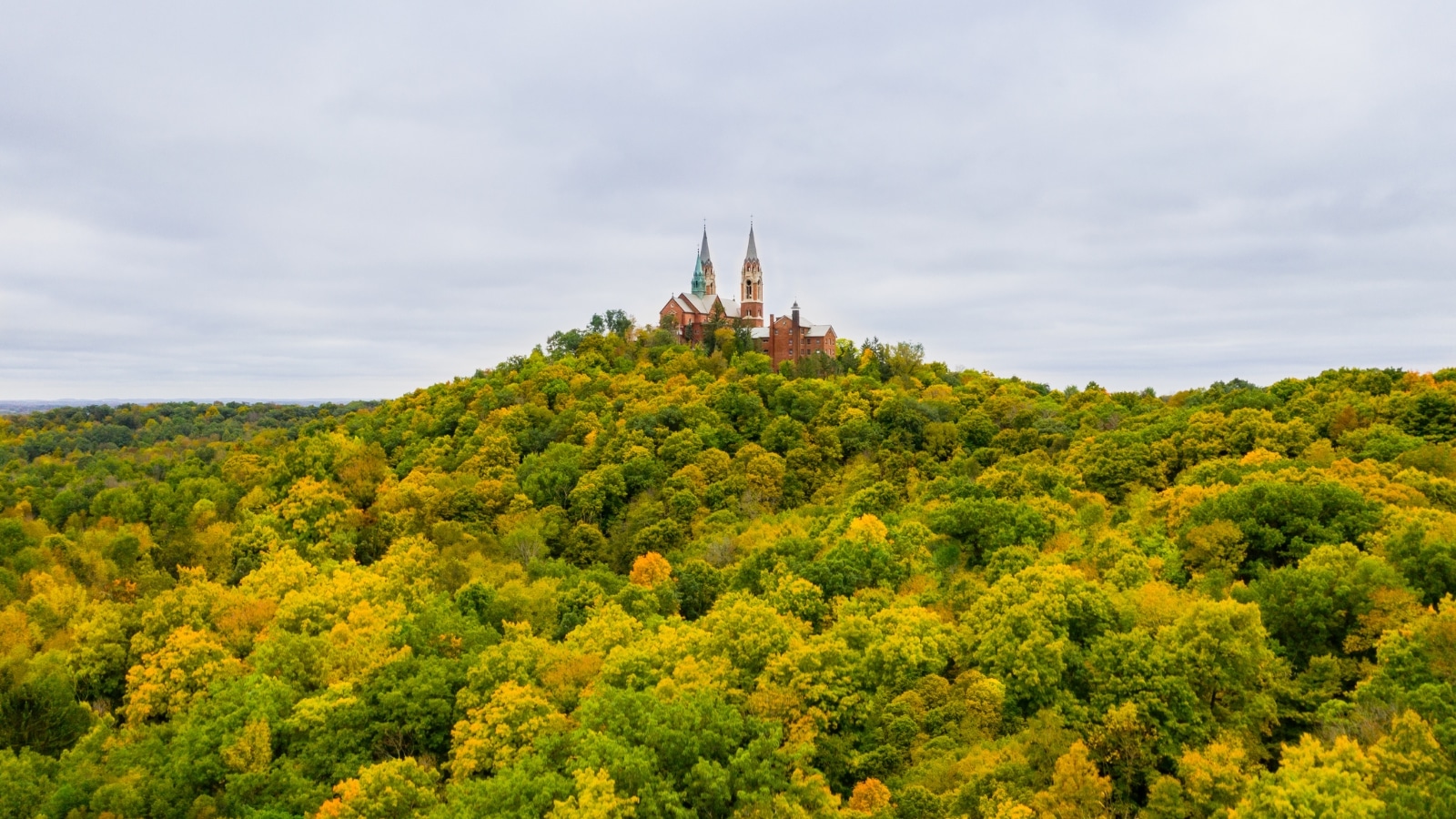 A view of the Holy Hill Basilica on a cloudy fall day. The cathedral sits atop a towering hill covered in lush trees, which are turning autumn colors of orange and green.