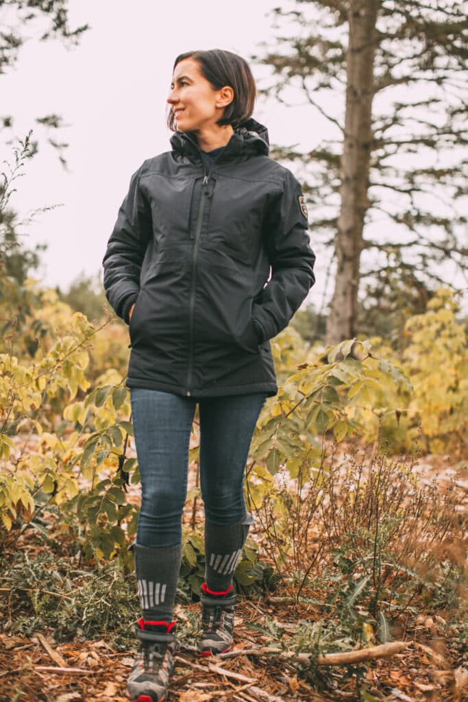 Lindsey of Have Clothes, Will Travel wearing a black United by Blue bison winter jacket in black with jeans and hiking boots in a wooded area.