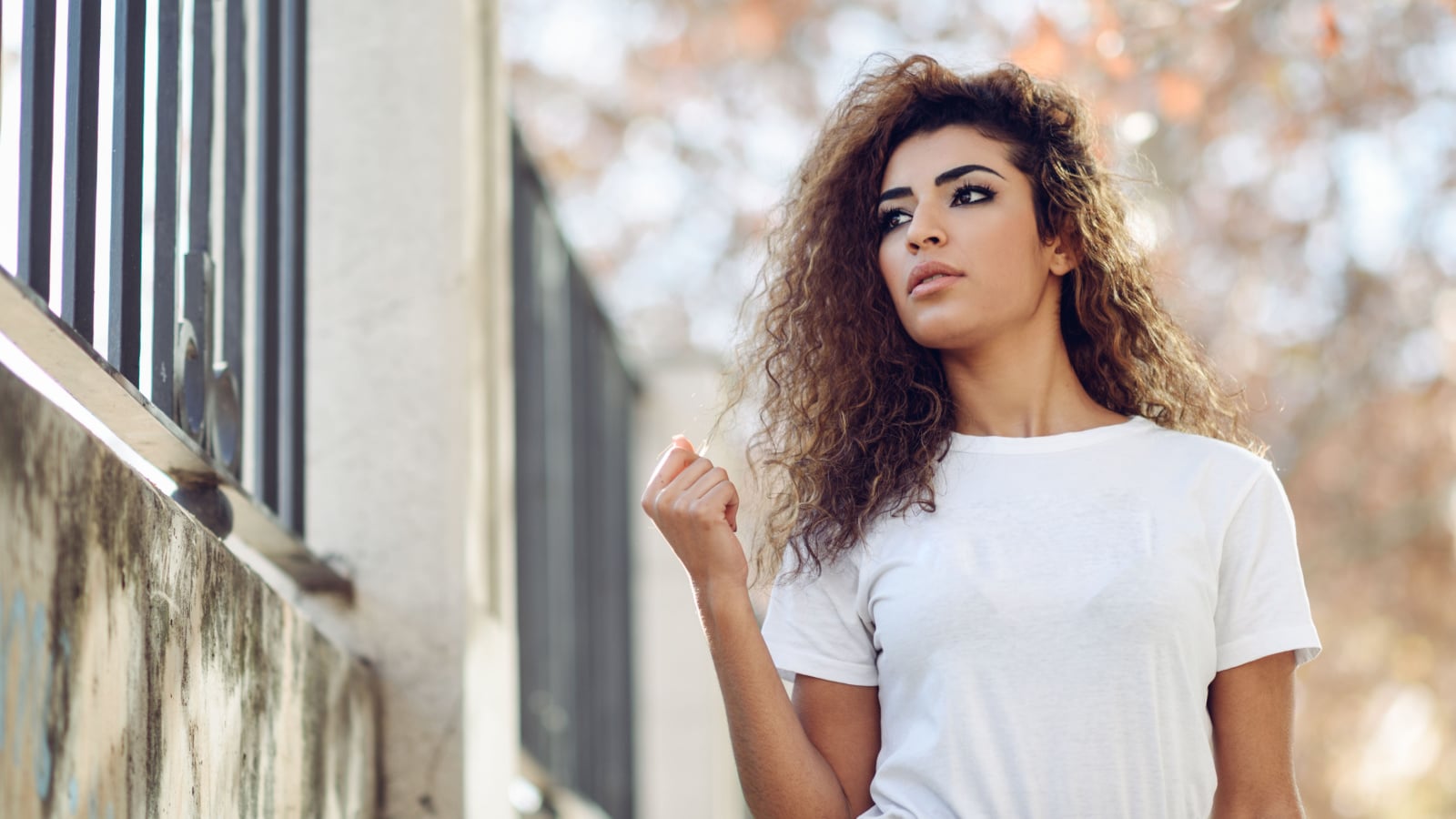 Beautiful young arabic woman with black curly hairstyle. Arab girl in casual clothes in the street. Happy female wearing white t-shirt and checked pants.