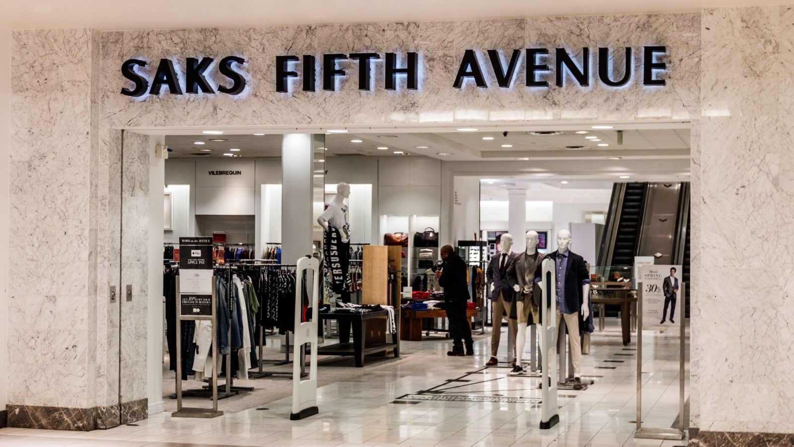 A storefront of Saks Fifth Avenue in a retail mall The storefront features the store name above the archway, mounted on a marble facade. Racks of clothing and mannequins fill the entrance of the store.