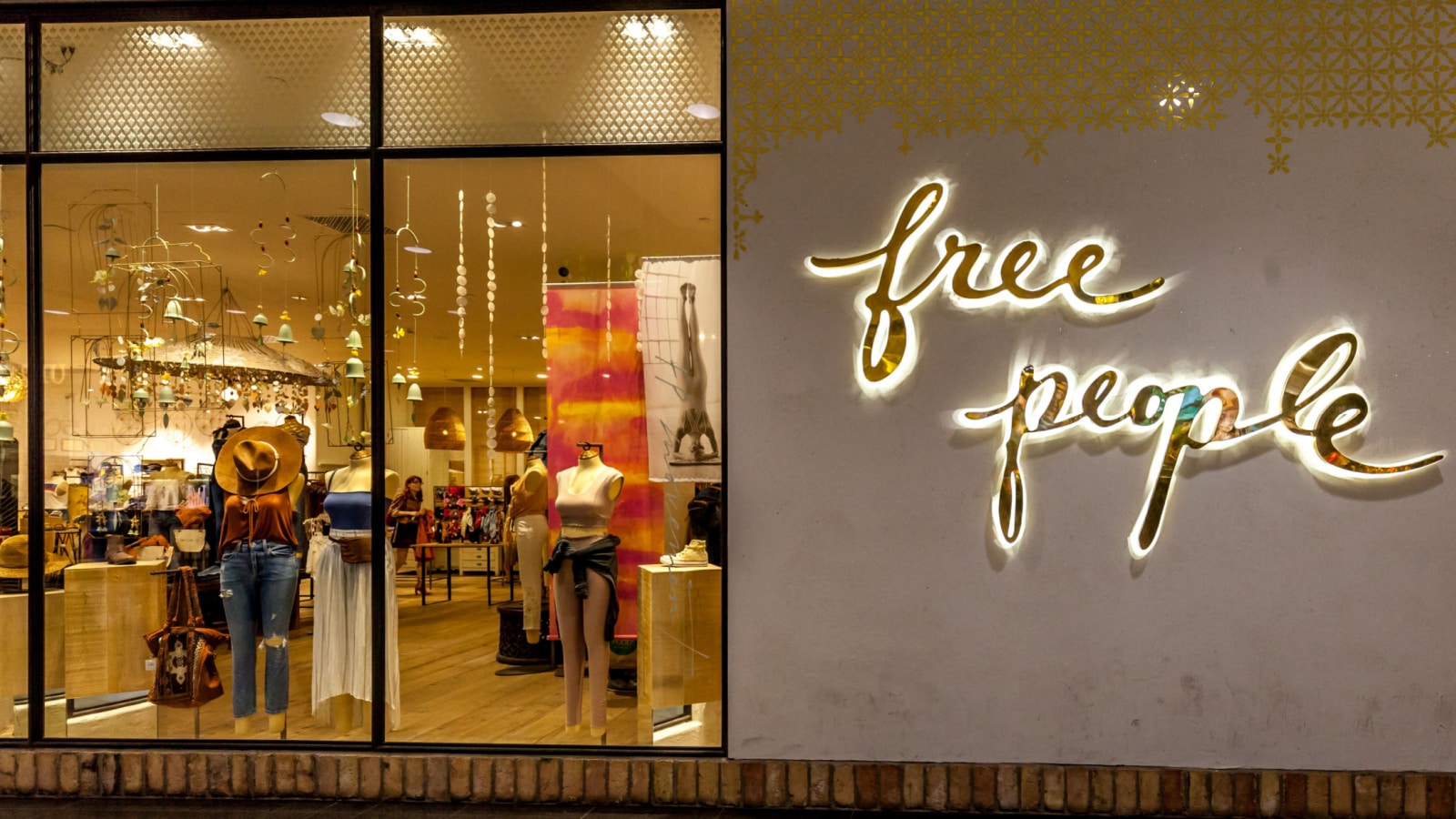 The storefront of Free People in a shopping mall. Large floor-to-ceiling windows look into the store at mannequins and clothing displays. The store name is mounted on the wall next to the windows, backlit by lights.