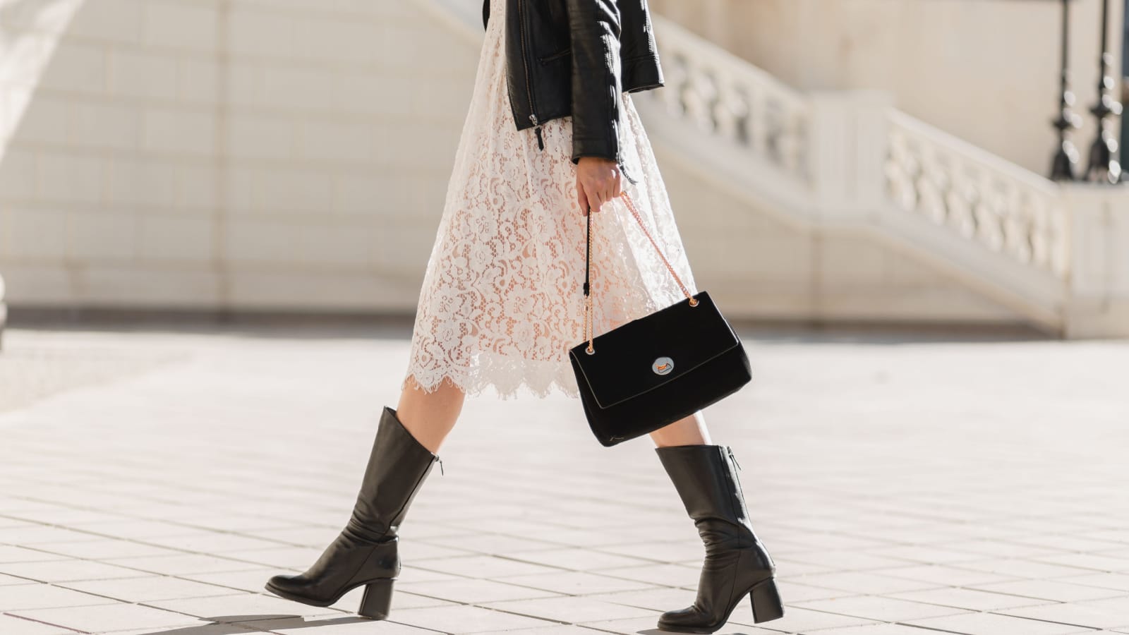 legs of attractive woman walking in street in high leather boots, fashionable outfit, holding purse, black leather jacket and white lace dress, spring autumn style, shoes fashion trend