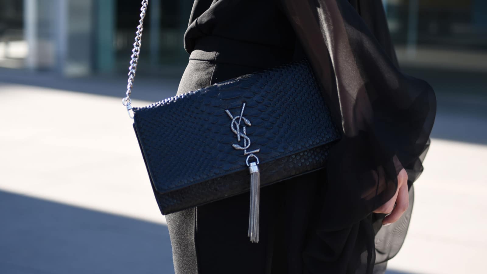 A closeup image of a woman, pictured from the waist down. She's wearing all black clothing and a designer YSL bag on her shoulder.