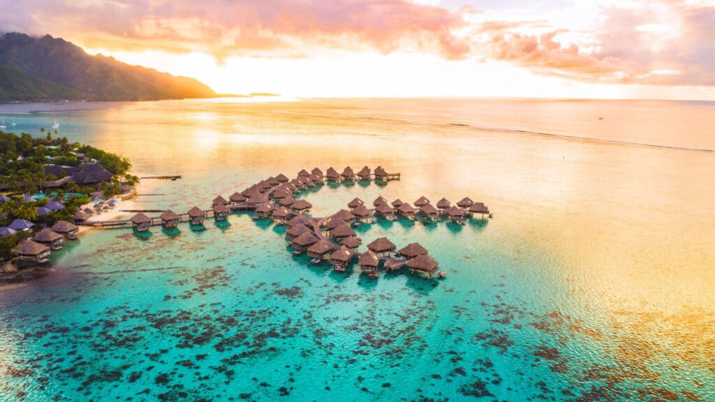 Luxury travel vacation aerial of overwater bungalows resort in coral reef lagoon ocean by beach. View from above at sunset of paradise getaway Moorea, French Polynesia, Tahiti, South Pacific Ocean.