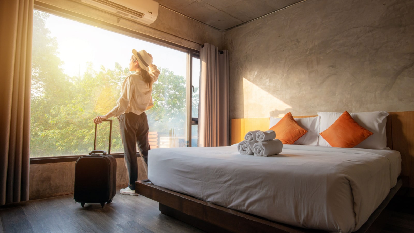 Portrait of tourist woman standing nearly window, looking to beautiful view with her luggage in hotel bedroom after check-in. Conceptual of travel and vacation.