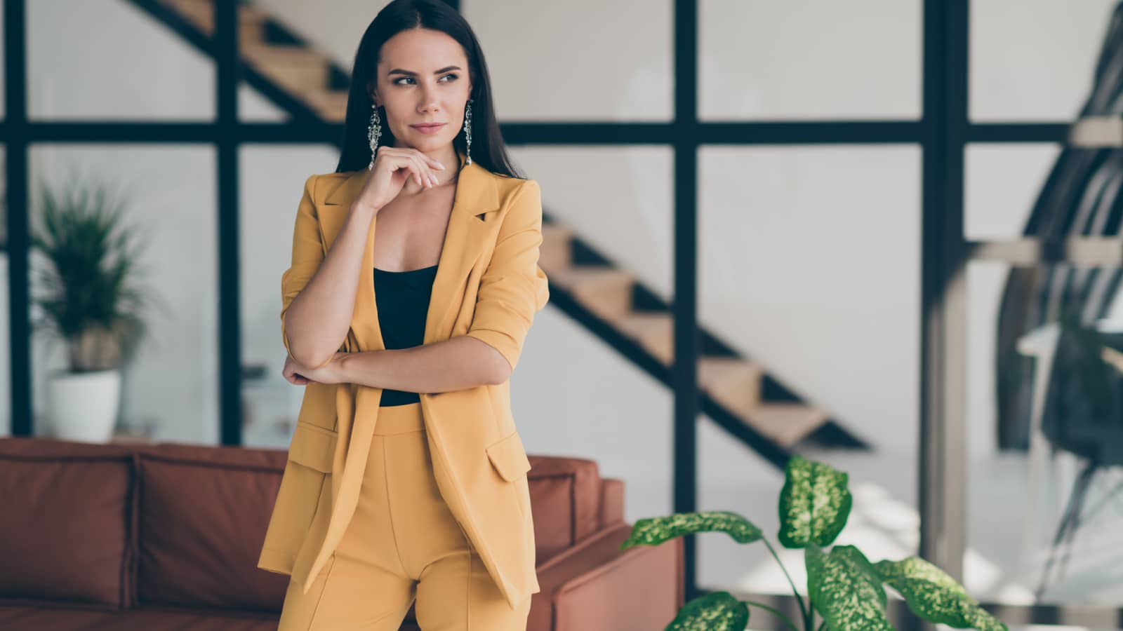 A picture of a young, stylish woman wearing a marigold yellow two piece suit over a black top. She's standing in a modern living room space, posed confidently, looking to her left.