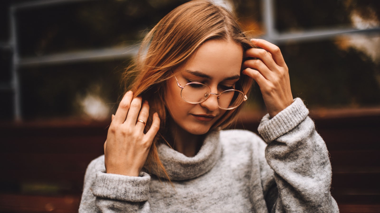 Young woman in a knitted grey turtleneck sweater fixed her hair. Close up. Blonde girl in glasses basking in grey turtleneck. Outdoor close up portrait of young beautiful woman.