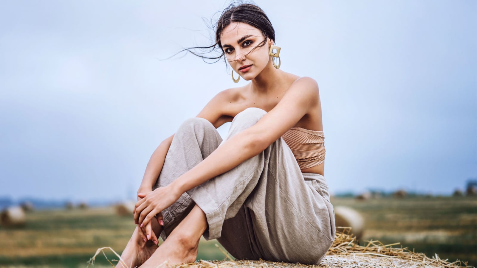 Brunette in linen pants and bare shoulders sitting on a hay bales in warm autumn day. Woman looking at camera. Behind her is a wheat field.