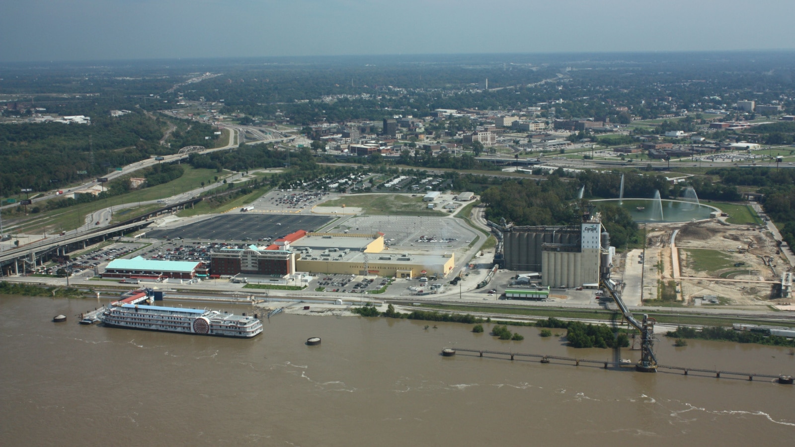 An aerial photo of East St. Louis on the Illinois side of the Mississippi River, showing a flat landscape with large parking lots, roads, and trees.