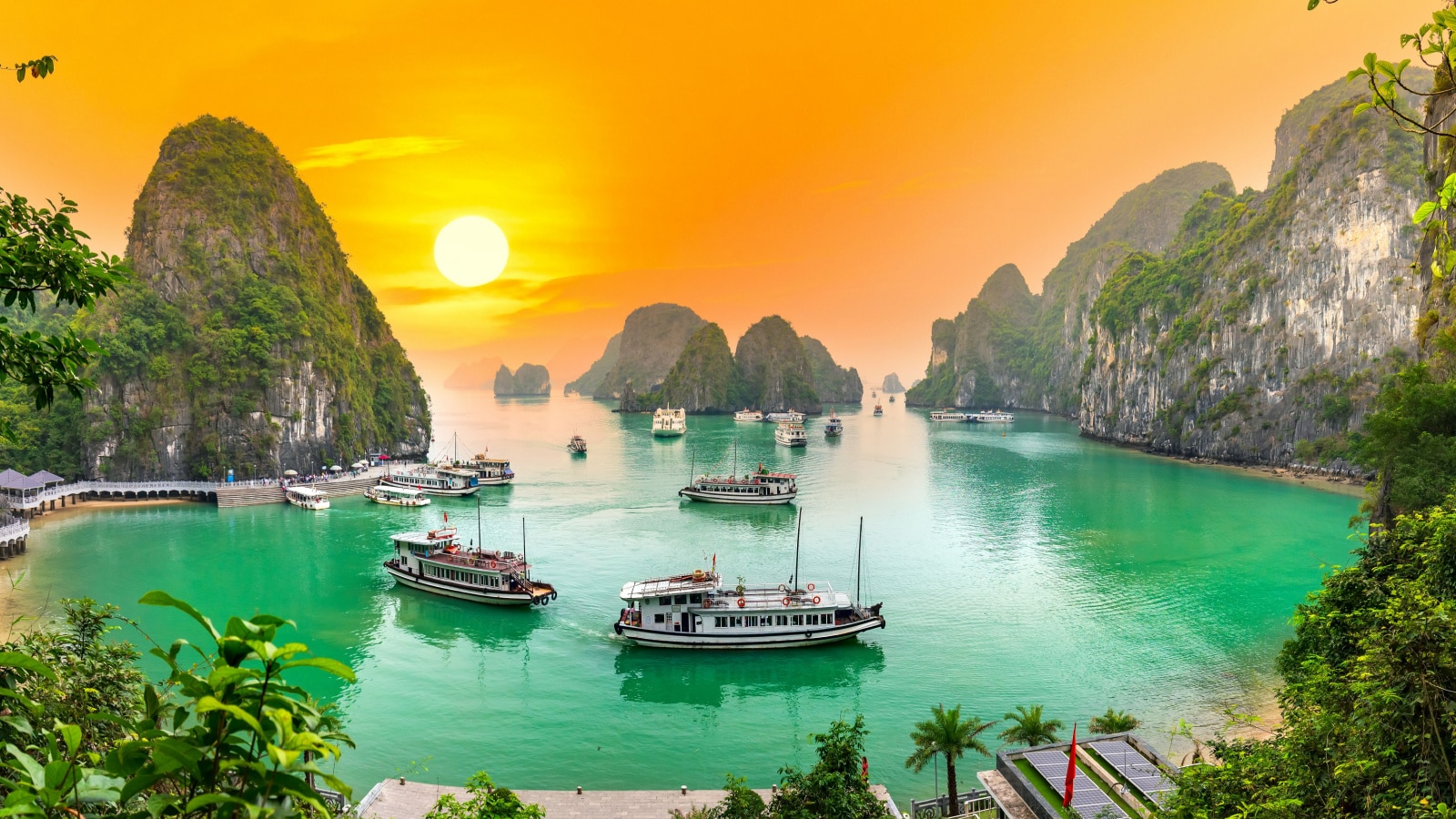 An image of a dreamy sunset landscape Halong Bay, Vietnam. The large sun fills the orange sky. Crystal blue waters fill the bay, where ships circle in and out of the bay waters.