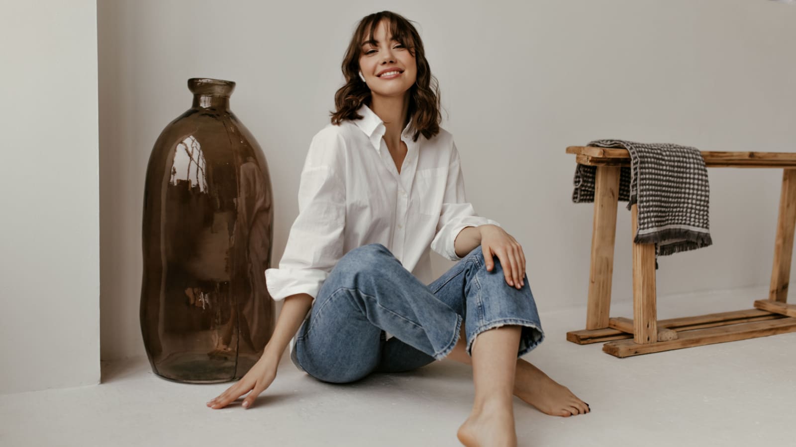 Joyful brunette curly woman in trendy pants and white shirt smiles and looks into camera. Pretty lady in jeans sits on floor in bathroom.