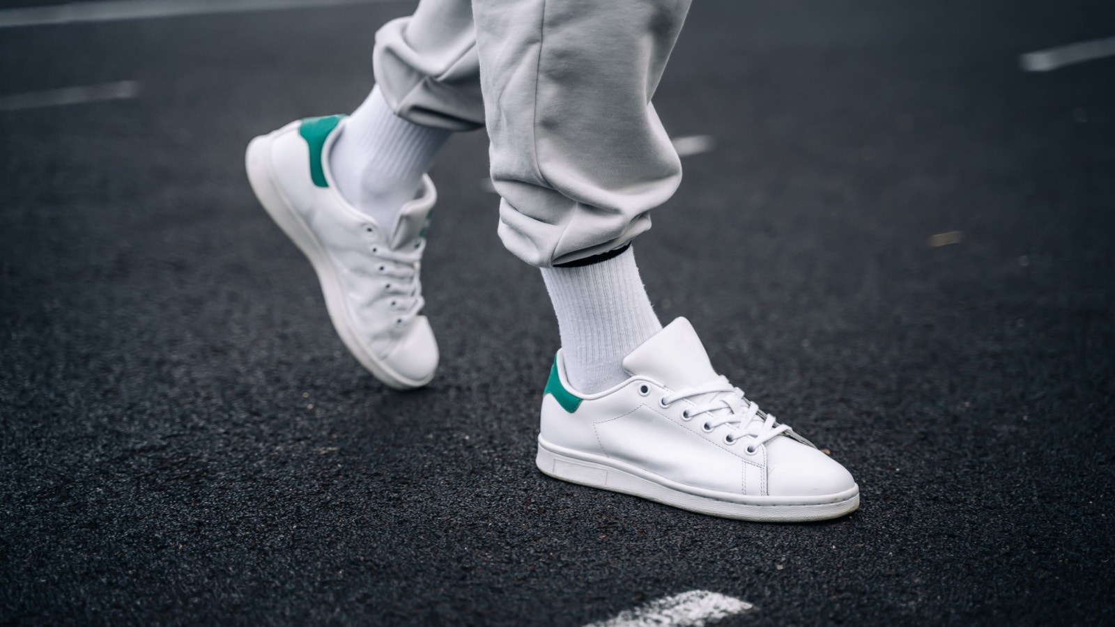 Women's legs in sweatpants, white socks and white sneakers with green elements against the background of the track with markings in the stadium.