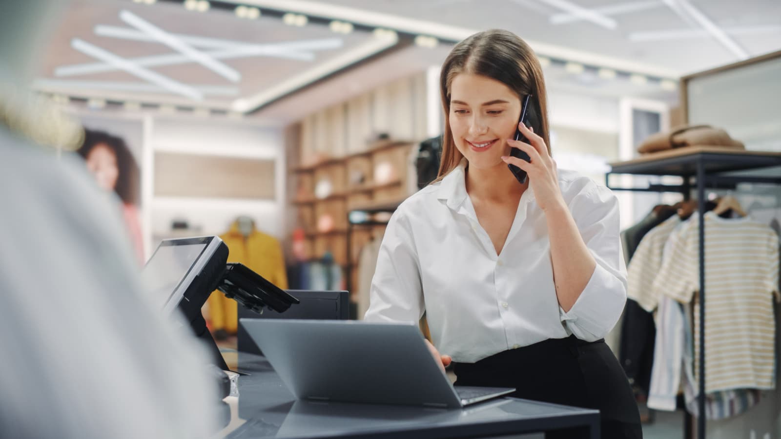 A clothing retail worker smiles while talking on a smartphone and working on her computer. She's standing at the checkout counter with clothing racks behind her.