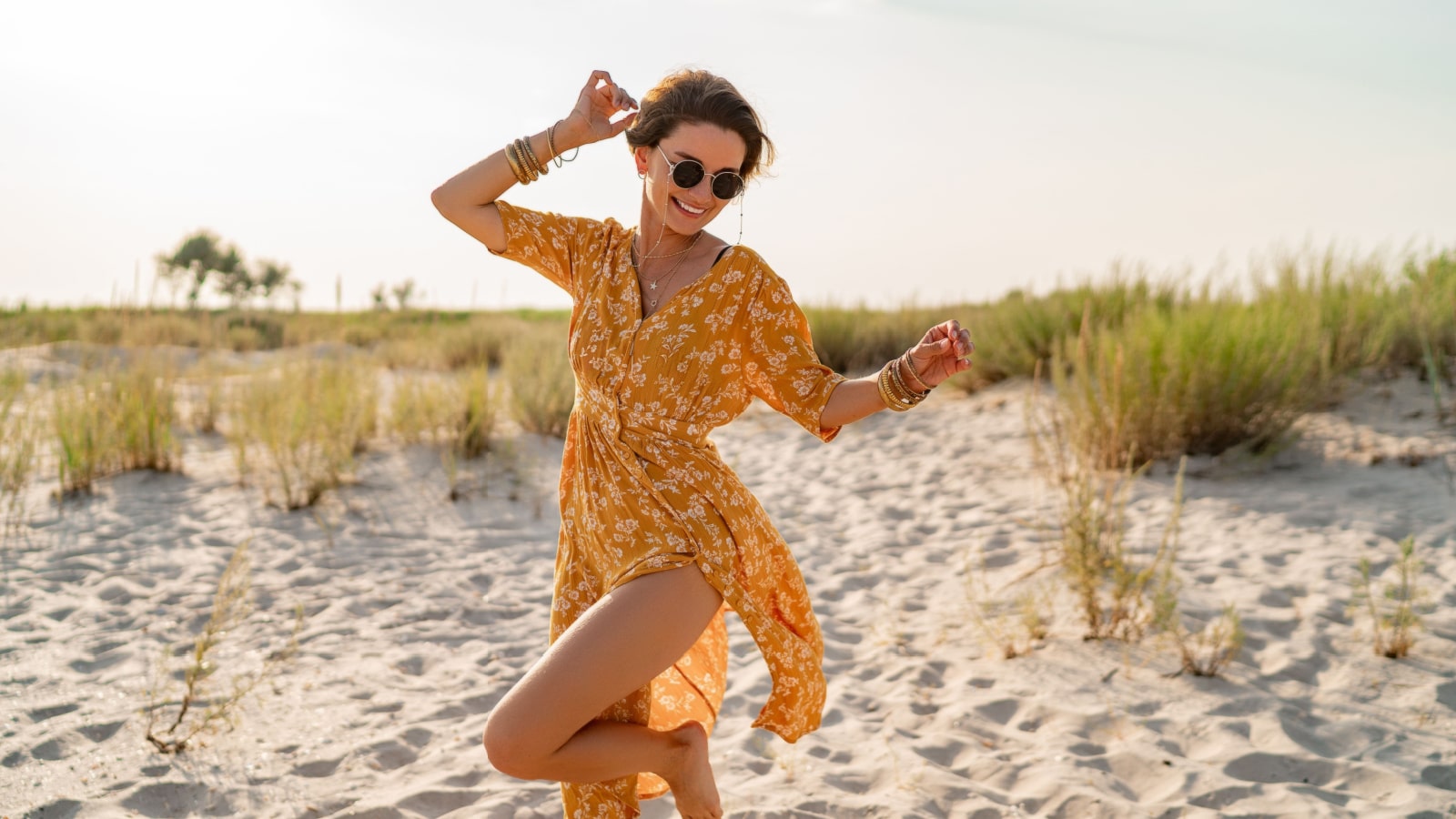 stylish attractive slim smiling woman on beach in summer style fashion trend outfit carefree and happy, feeling freedom, wearing yellow printed dress boho style chic and sunglasses