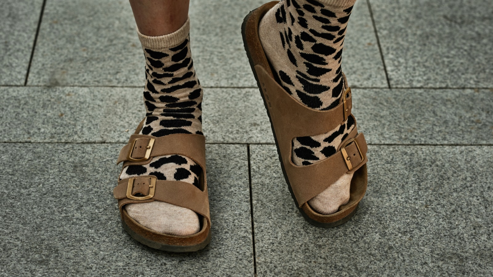 Female legs in beige leopard socks and brown sandals on background of paving slabs. European casual fashion.