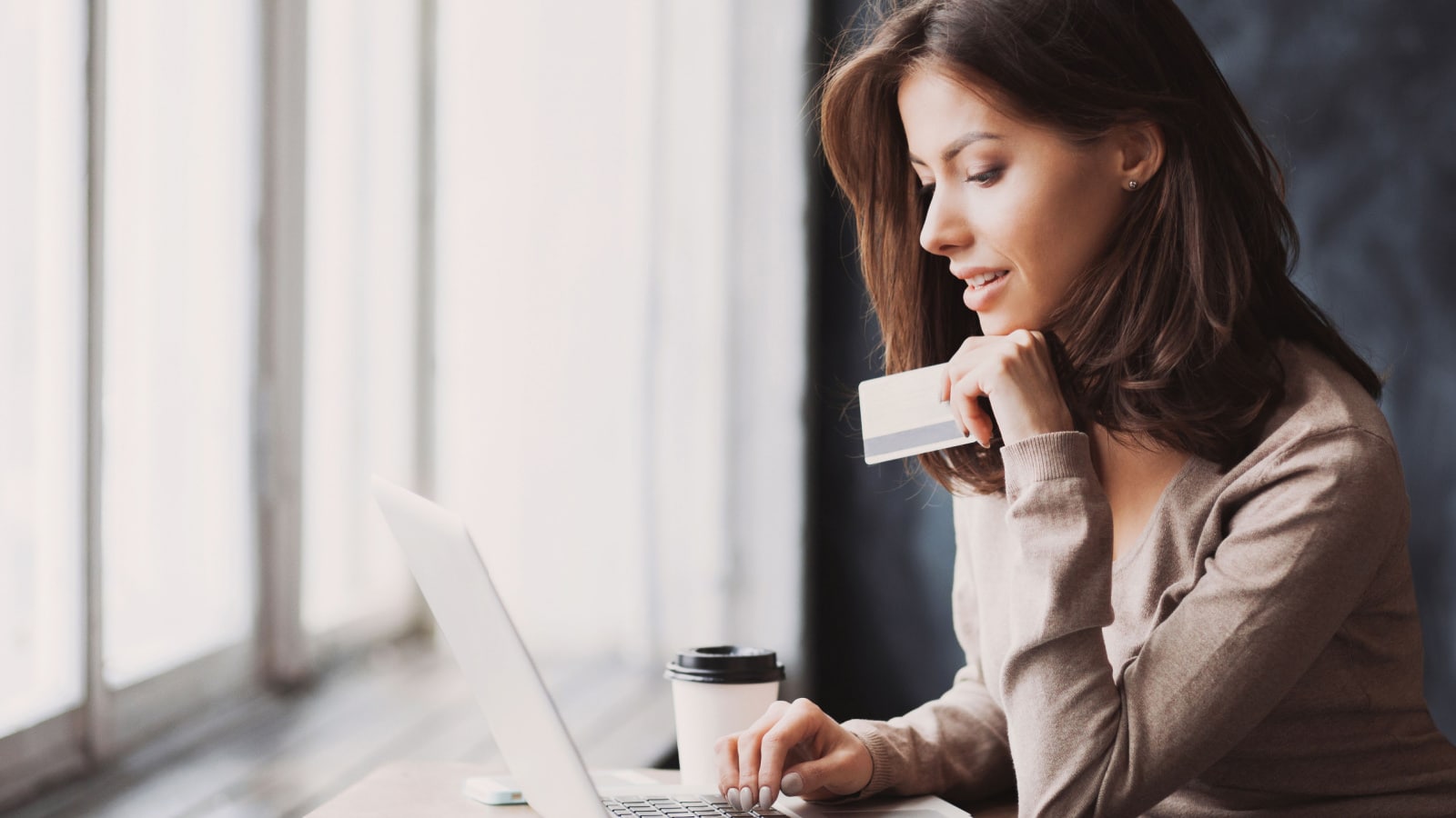 Young woman holding credit card and using laptop computer. She is sitting at a counter in front of large windows, with a to-go coffee cup next to her computer.