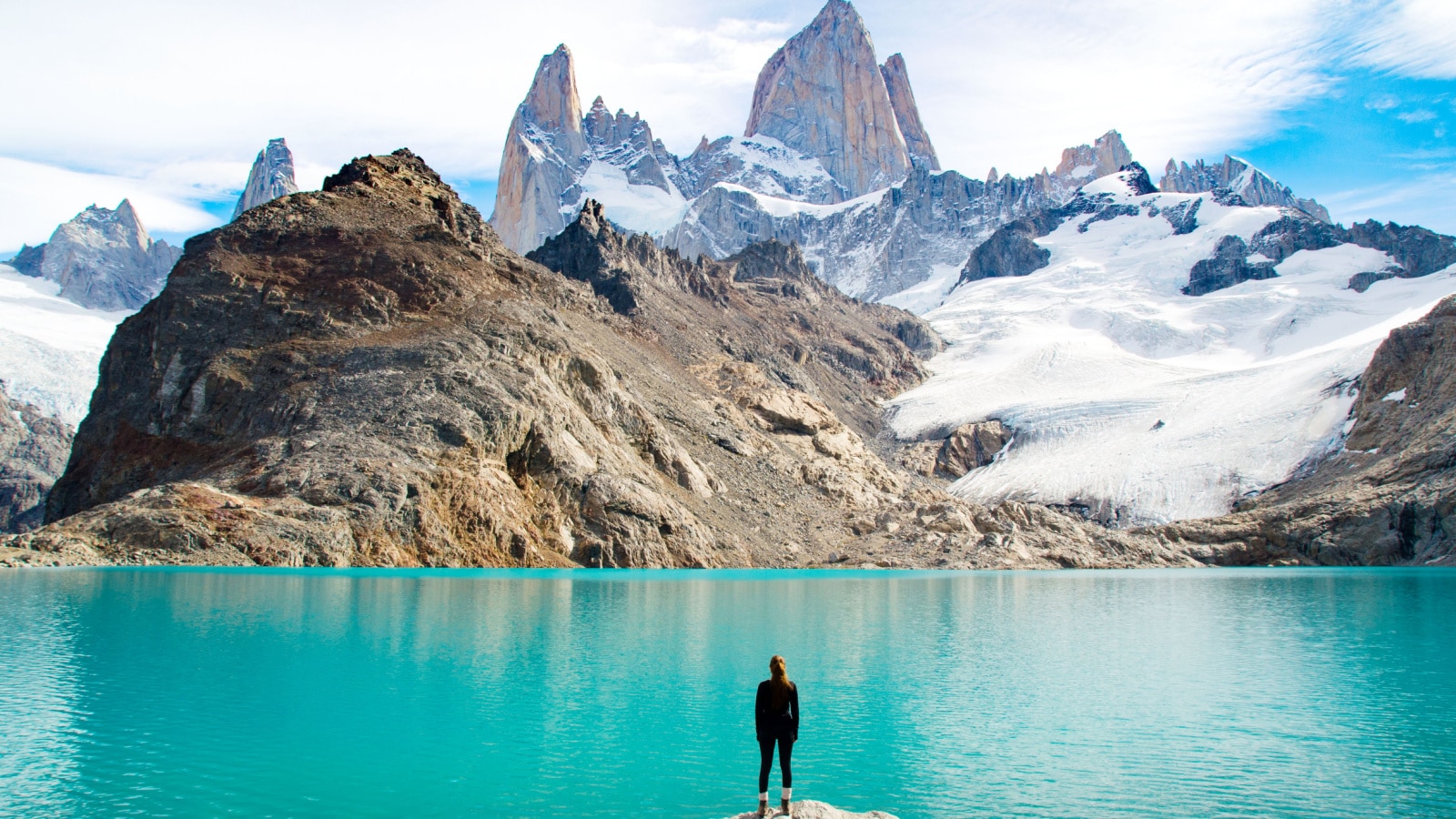 A woman stands at the shoreline of a clear blue lake at the base of Fitz Roy Mountain in Patagonia, looking at the stunning snow-covered mountain range range in front of her.