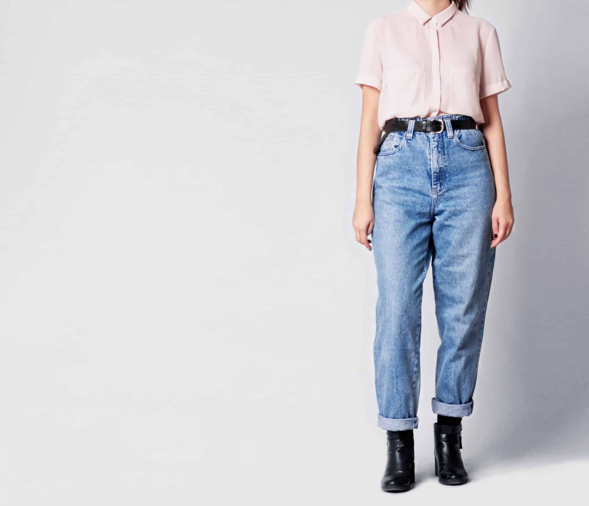 Woman wearing casual outfit with white shirt, blue vintage high-waisted mom jeans with black ankle boots and black belt isolated on grey background.