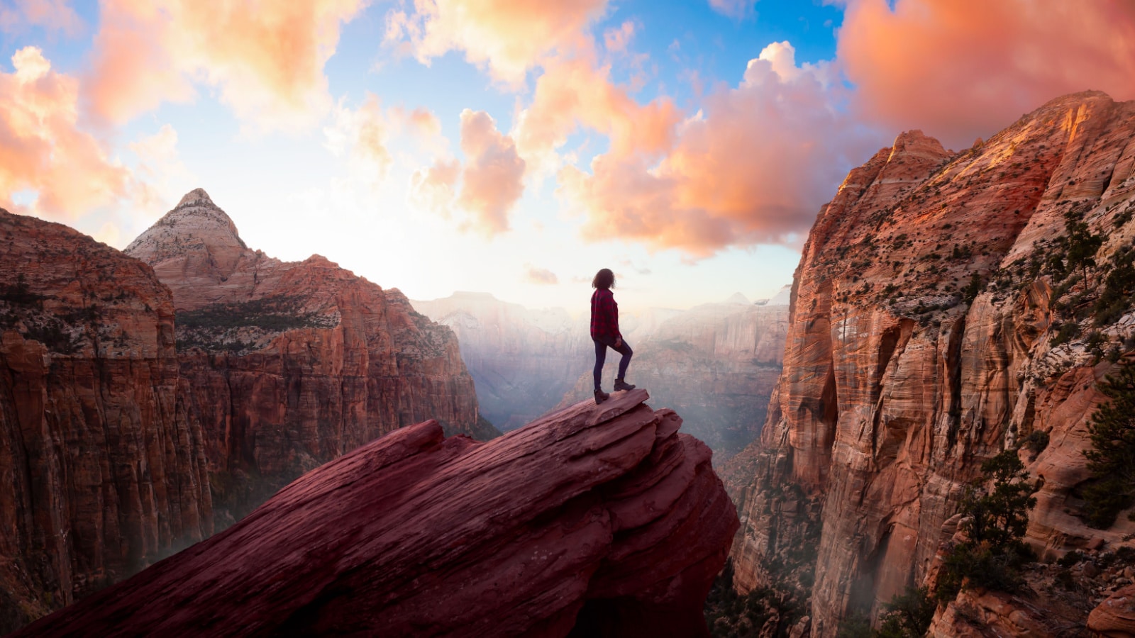 Adventurous Woman at the edge of a cliff is looking at a beautiful landscape view in the Canyon during a vibrant sunset. Taken in Zion National Park, Utah, United States. Sky Composite Panorama