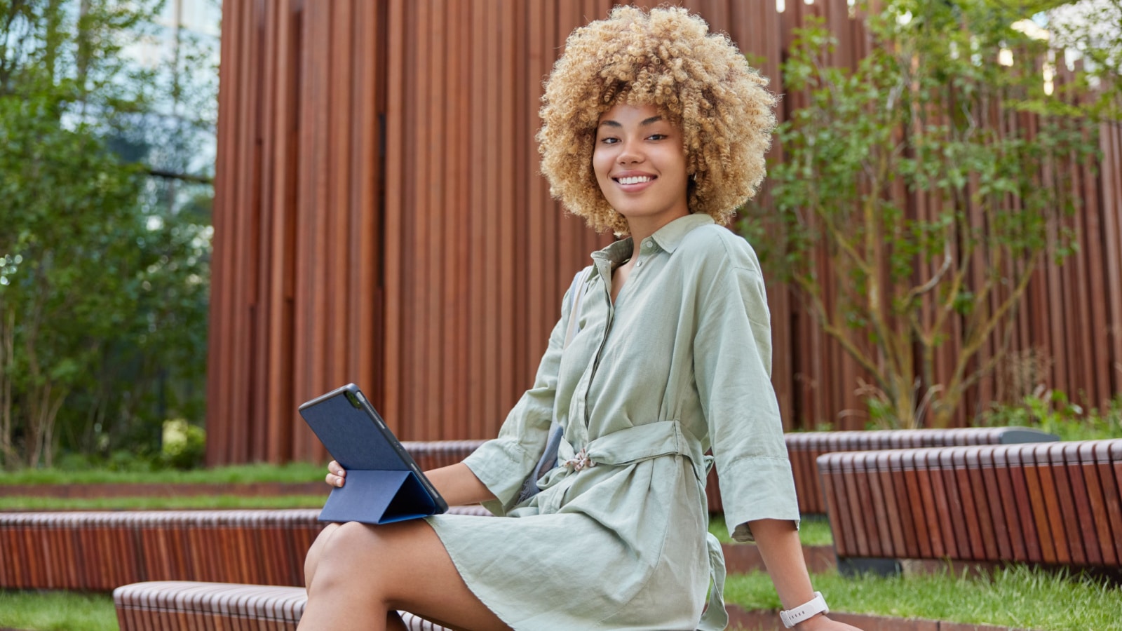 A smiling young woman sits on a park bench, holding a tablet in her lap, smiling at the camera.