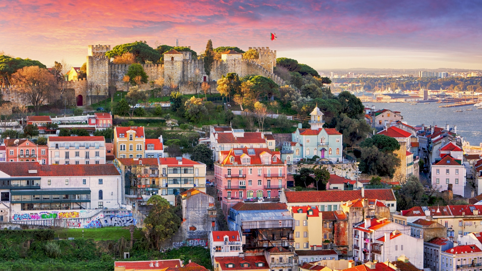 The colorful skyline of Lisbon, Portugal, featuring the Sao Jorge Castle at the top of the hill at dusk. Colorful buildings line the hillside, surrounded by trees and walkable city streets.