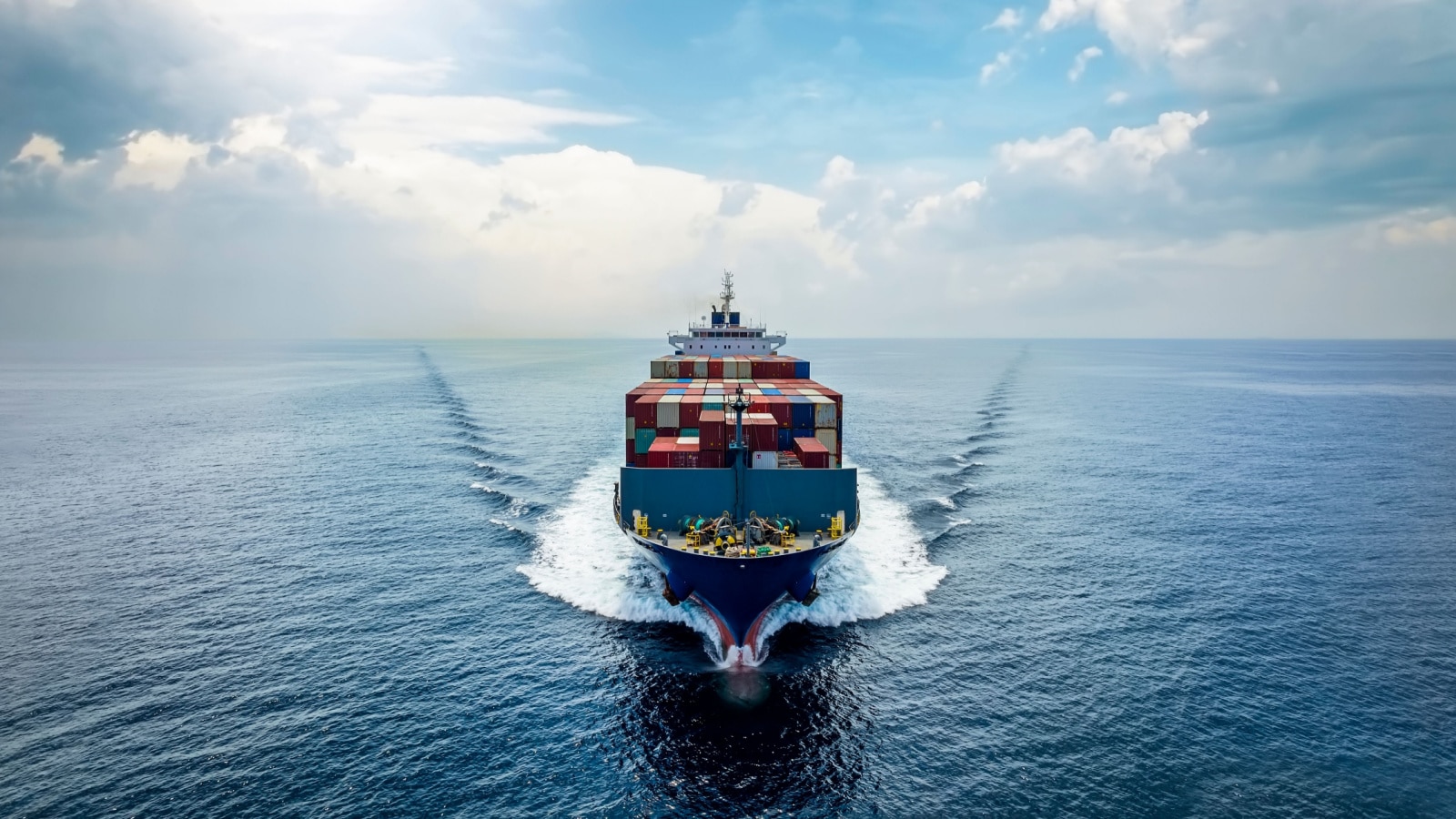 Aerial front view of a medium sized, loaded container cargo vessel traveling with speed over the ocean under cloudy sky