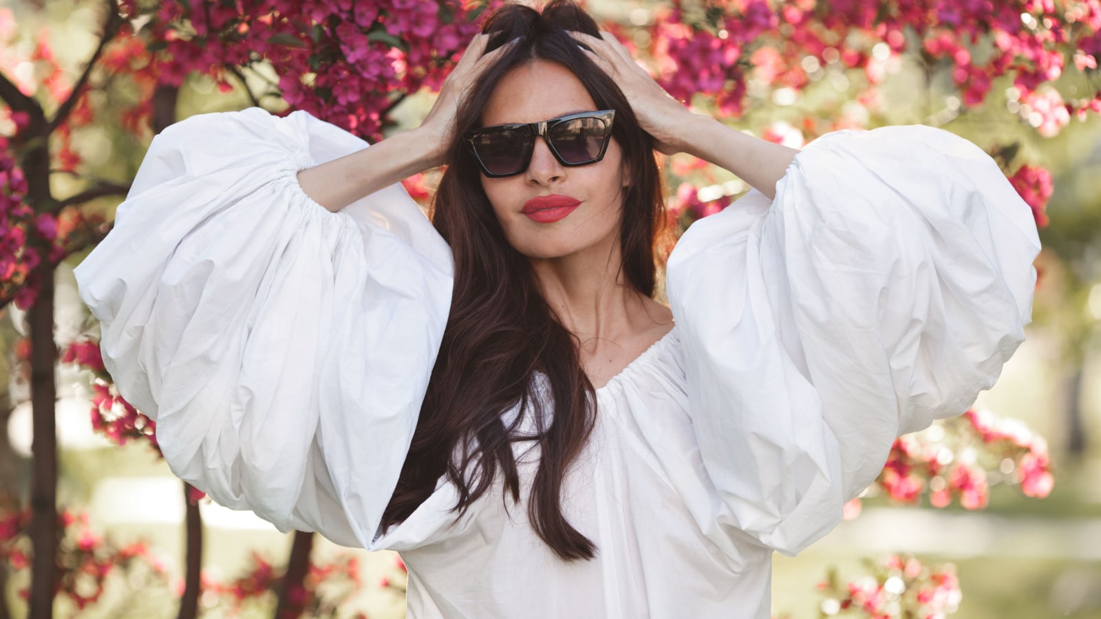 Gorgeous beautiful brunette woman with long hair in white cotton shirt with wide sleeves lanterns flounces on spring sakura blossoms background. Model touches hair