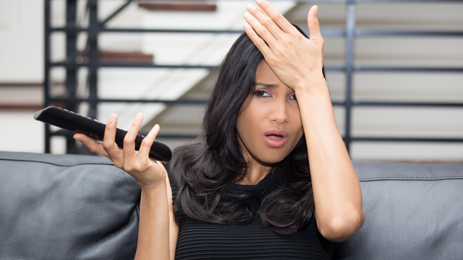 Closeup portrait, young woman sitting in black leather couch upset by what she sees on TV, isolated indoor flat background