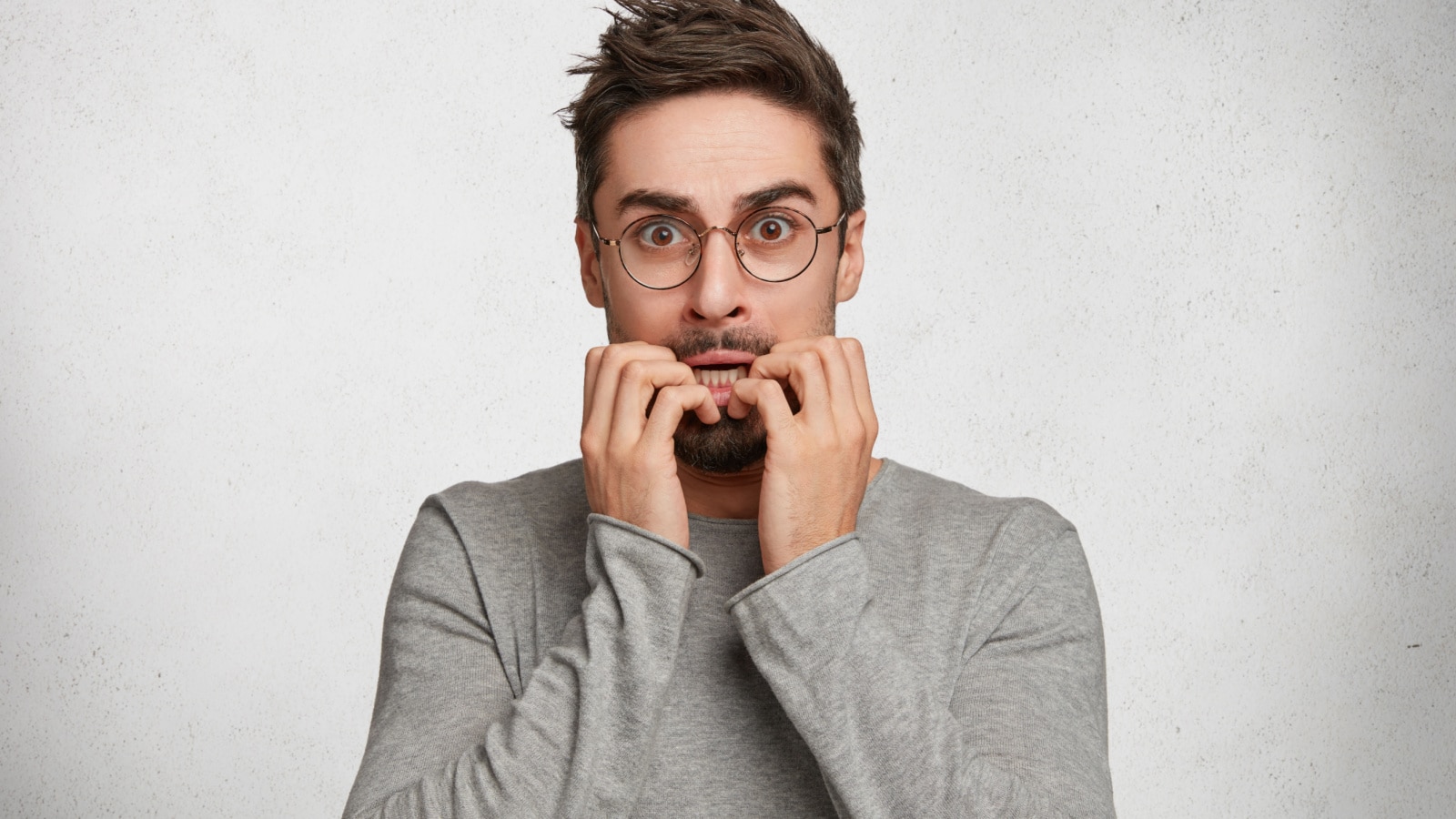 Indoor shot of nervous worried handsome man bites fingers, being scared of horror film or afraid of exams, has frightened expression, isolated over white concrete background. Emotional adult